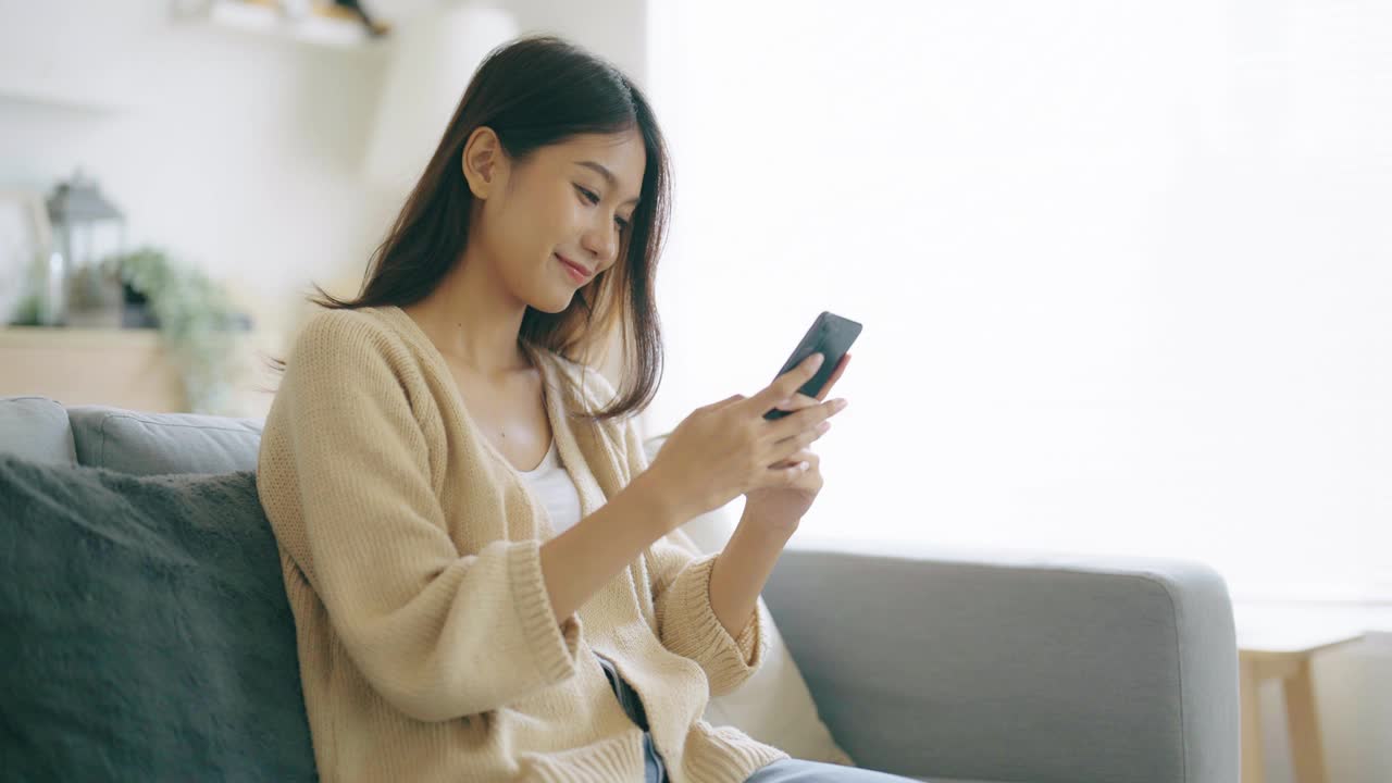 快乐的年轻亚洲女人在家里舒适的沙发上放松，在智能手机上输入聊天信息，微笑的女孩用手机聊天，在浏览器上搜索信息，无线互联网，网上购物视频素材