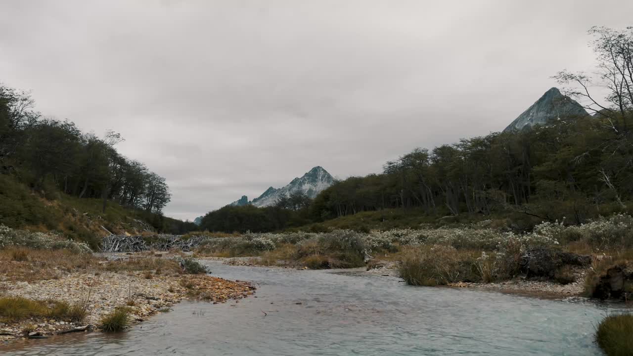 在阿根廷火地岛的乌斯怀亚，一名男子坐在河岸上，欣赏森林和山脉的美丽。-广角镜头视频素材