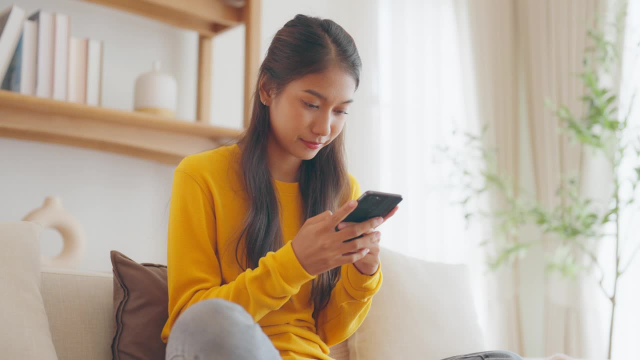 快乐的年轻亚洲女人放松在家里舒适的沙发上发短信在智能手机上，微笑的女孩使用手机，聊天在线消息，从家里网上购物视频素材