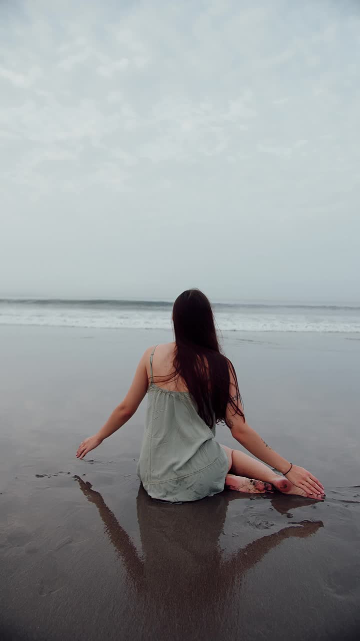 穿着裙子的女孩坐在海浪背景下的黑色沙滩上视频素材