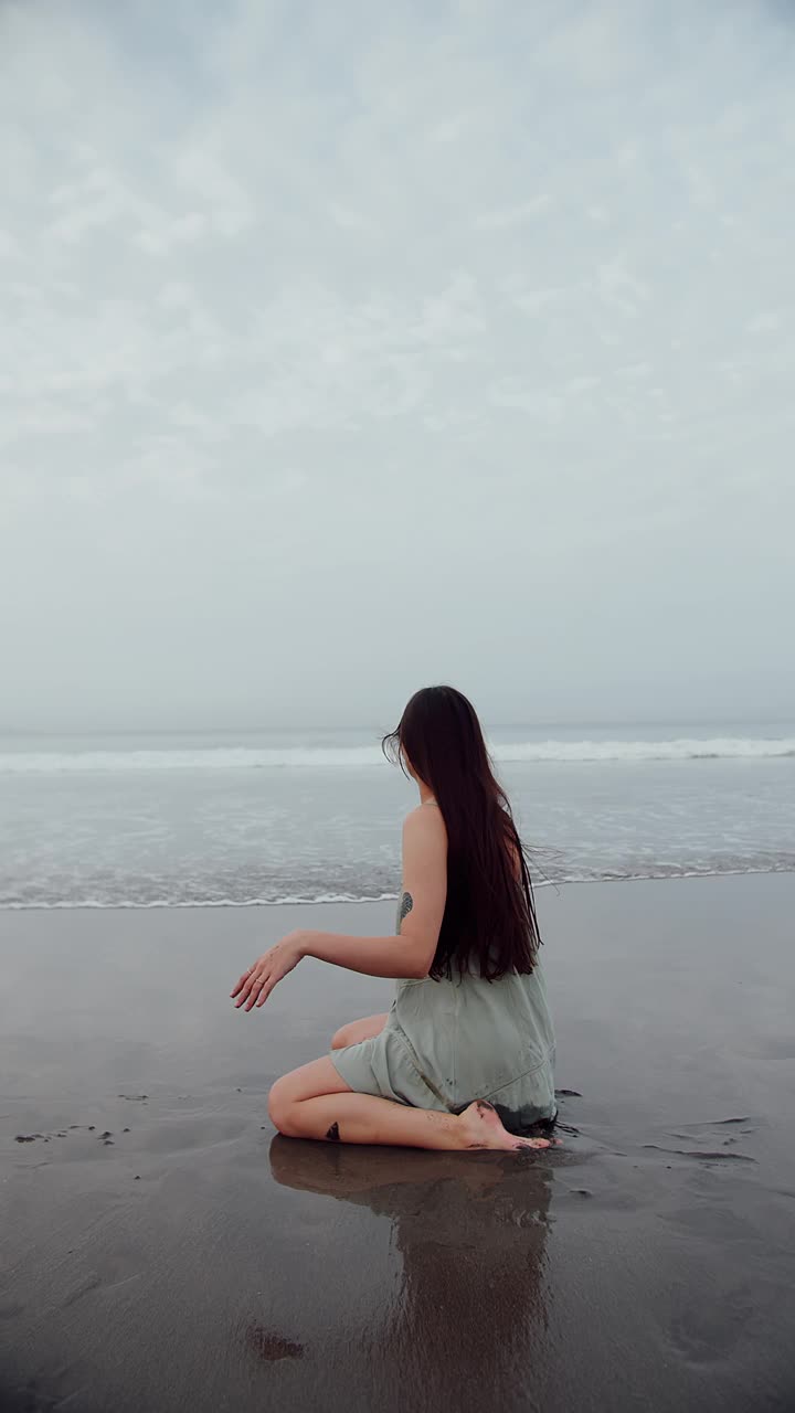 女孩坐在水里的海洋上，黑色的沙滩上有波浪。视频素材