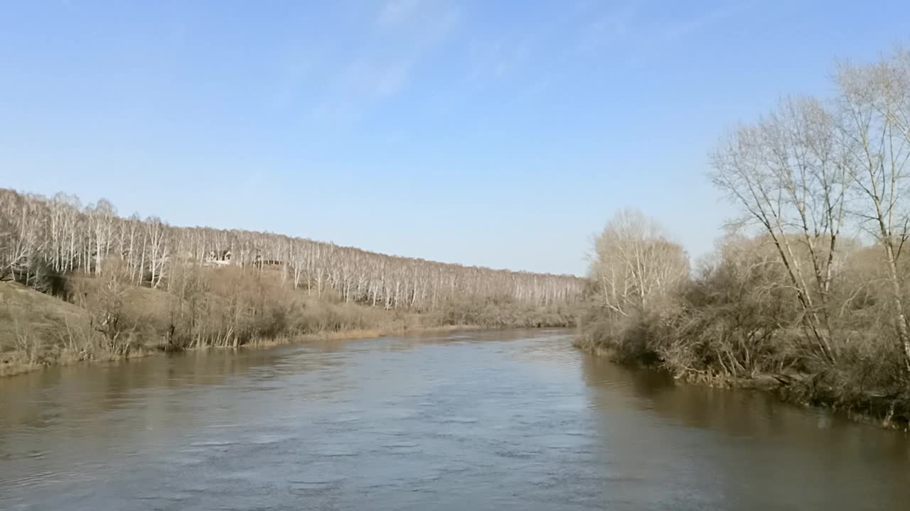 在一个晴朗的春日傍晚，一条平静的小河流经山谷，河床的拐弯处有几棵高大的光秃秃的树。视频素材