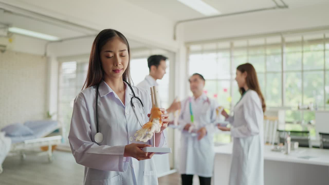 穿着制服的年轻亚洲女性骨科医生的肖像，抱着人体踝关节骨骼模型，在医院对着镜头微笑。医疗整形外科医师视频下载