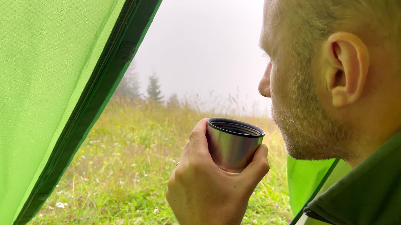 男子在帐篷里喝着热茶，在山谷营地享受秋雨的早晨。活跃人物活动和旅行概念4K素材。视频素材