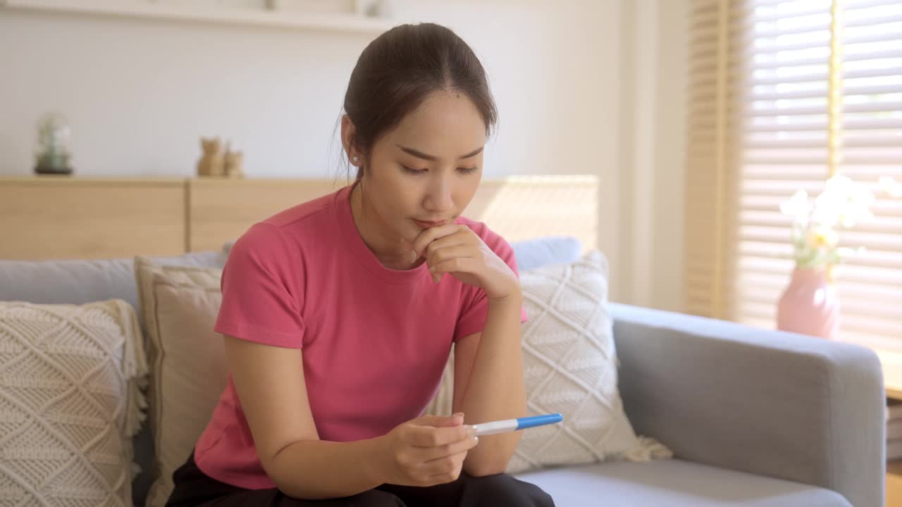 一位年轻的亚洲妇女在做了验孕棒后，坐在家里的沙发上，感到不高兴和焦虑。视频素材