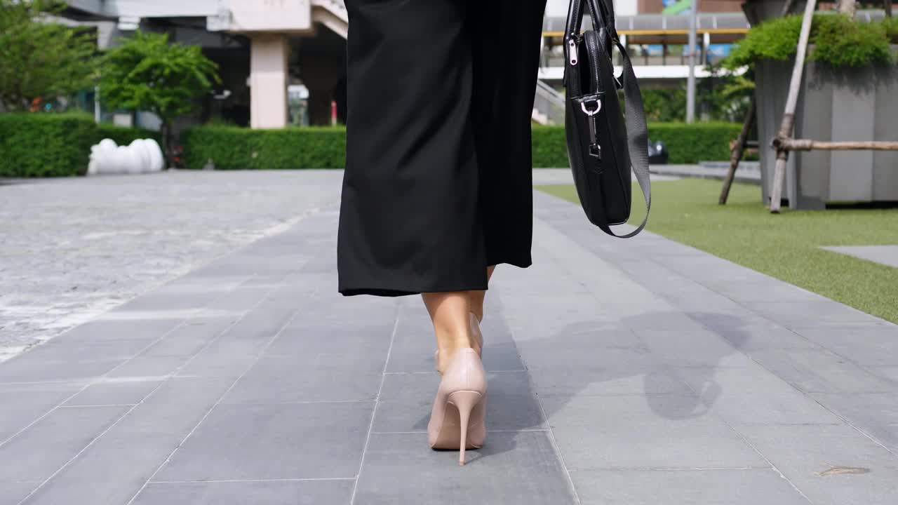 在一个温暖的夏日，一个女人穿着高跟鞋，手里拿着包走在城市里。这个女人心情很好，这从她模特的步态就可以看出来。女人自信地走在柏油路上去上班。视频下载