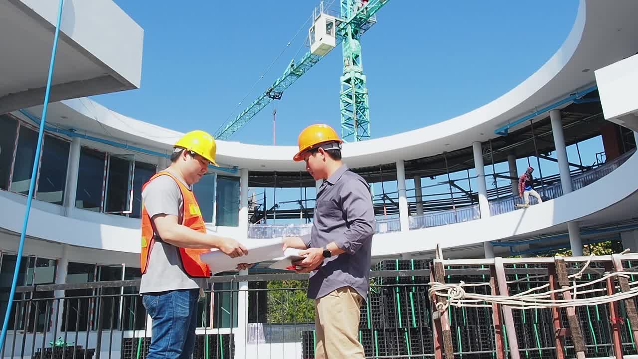 两名年轻的结构工程师和建筑师穿着橙色的工作背心站在机械起重机前施工现场。视频下载