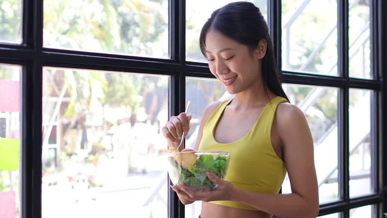 美丽健康的亚洲女人愉快地在家里吃美味的蔬菜沙拉。穿着运动服的漂亮运动女孩喜欢在运动后吃干净的蔬菜，在家里保持健康。饮食和健康食品。视频素材