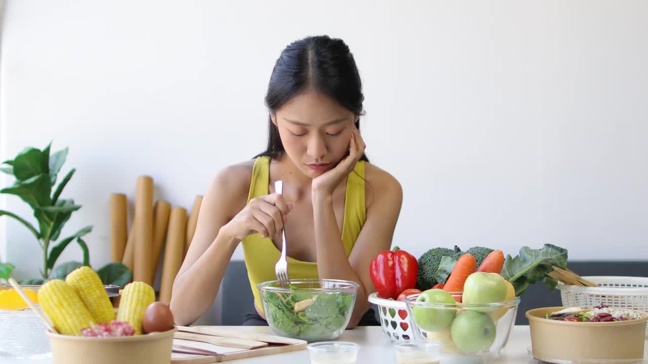 一位每天吃蔬菜沙拉的年轻亚洲女性对蔬菜感到厌倦。不快乐的女人在节食的时候看着叉子上的蔬菜。女孩不想吃蔬菜，不喜欢味道。视频素材