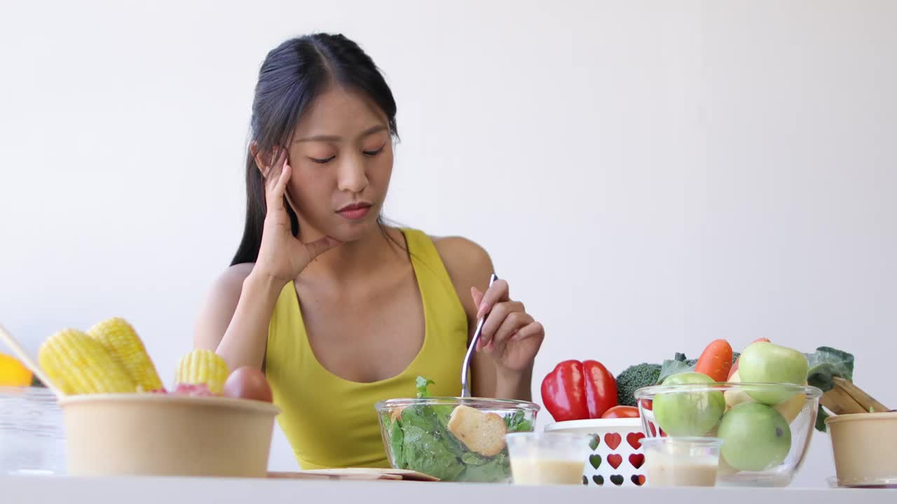 一位每天吃蔬菜沙拉的年轻亚洲女性对蔬菜感到厌倦。不快乐的女人在节食的时候看着叉子上的蔬菜。女孩不想吃蔬菜，不喜欢味道。视频素材