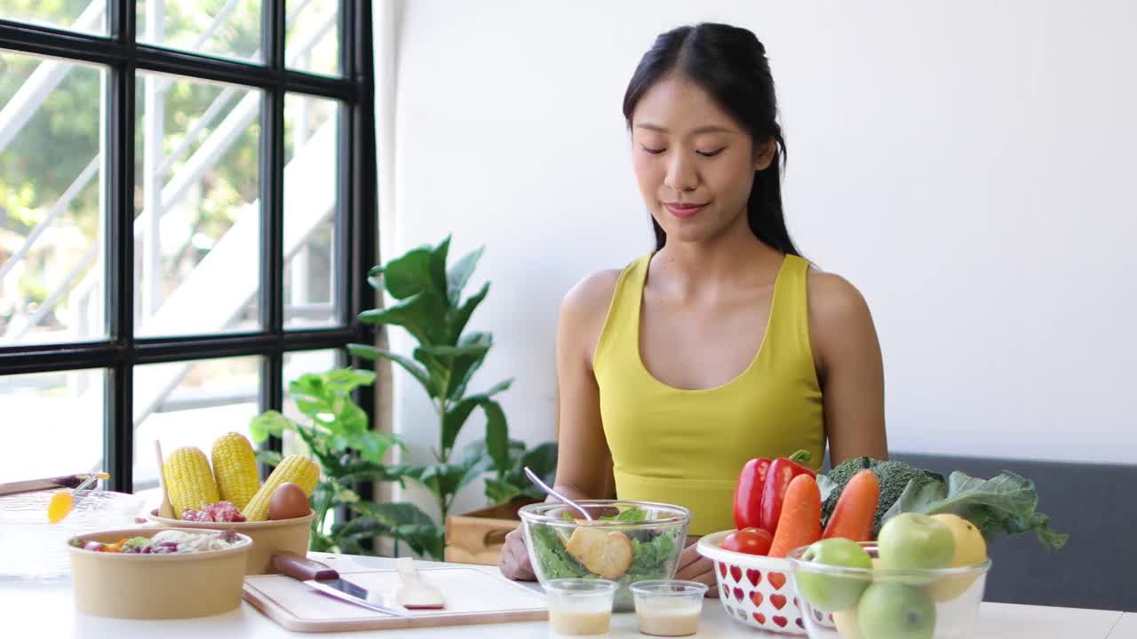 美丽健康的亚洲女人愉快地在家里吃美味的蔬菜沙拉。穿着运动服的漂亮运动女孩喜欢在运动后吃干净的蔬菜，在家里保持健康。饮食和健康食品。视频素材