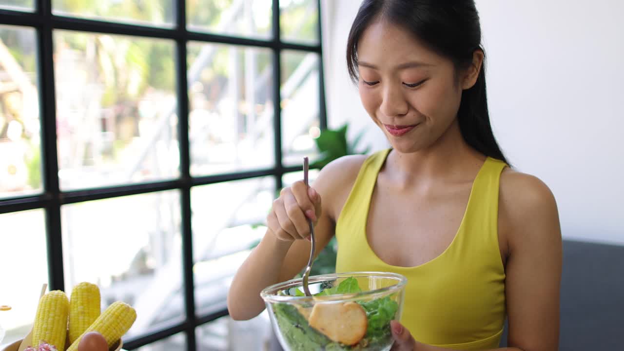 美丽健康的亚洲女人愉快地在家里吃美味的蔬菜沙拉。穿着运动服的漂亮运动女孩喜欢在运动后吃干净的蔬菜，在家里保持健康。饮食和健康食品。视频素材