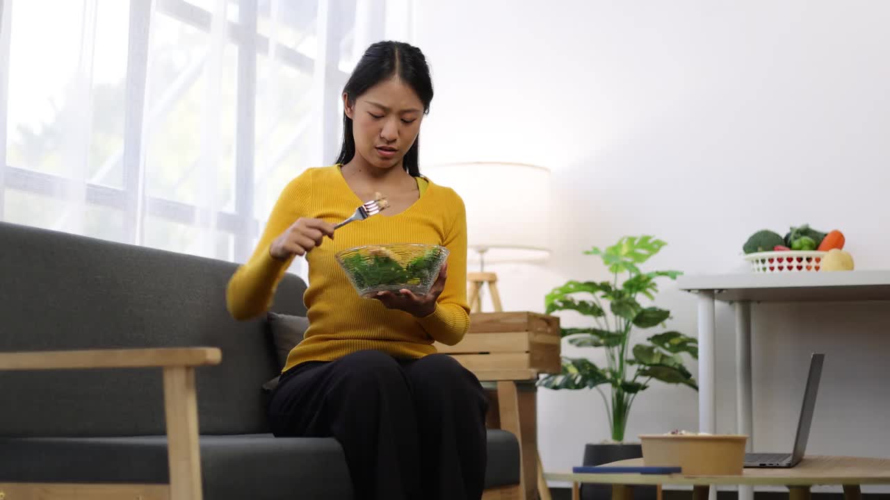 一位每天吃蔬菜沙拉的年轻亚洲女性对蔬菜感到厌倦。不快乐的女人在节食的时候看着叉子上的蔬菜。女孩不想吃蔬菜，不喜欢味道。视频素材