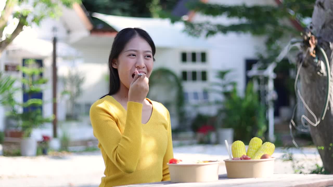 快乐的年轻亚洲女人在后院的木桌上吃蔬菜沙拉。视频素材