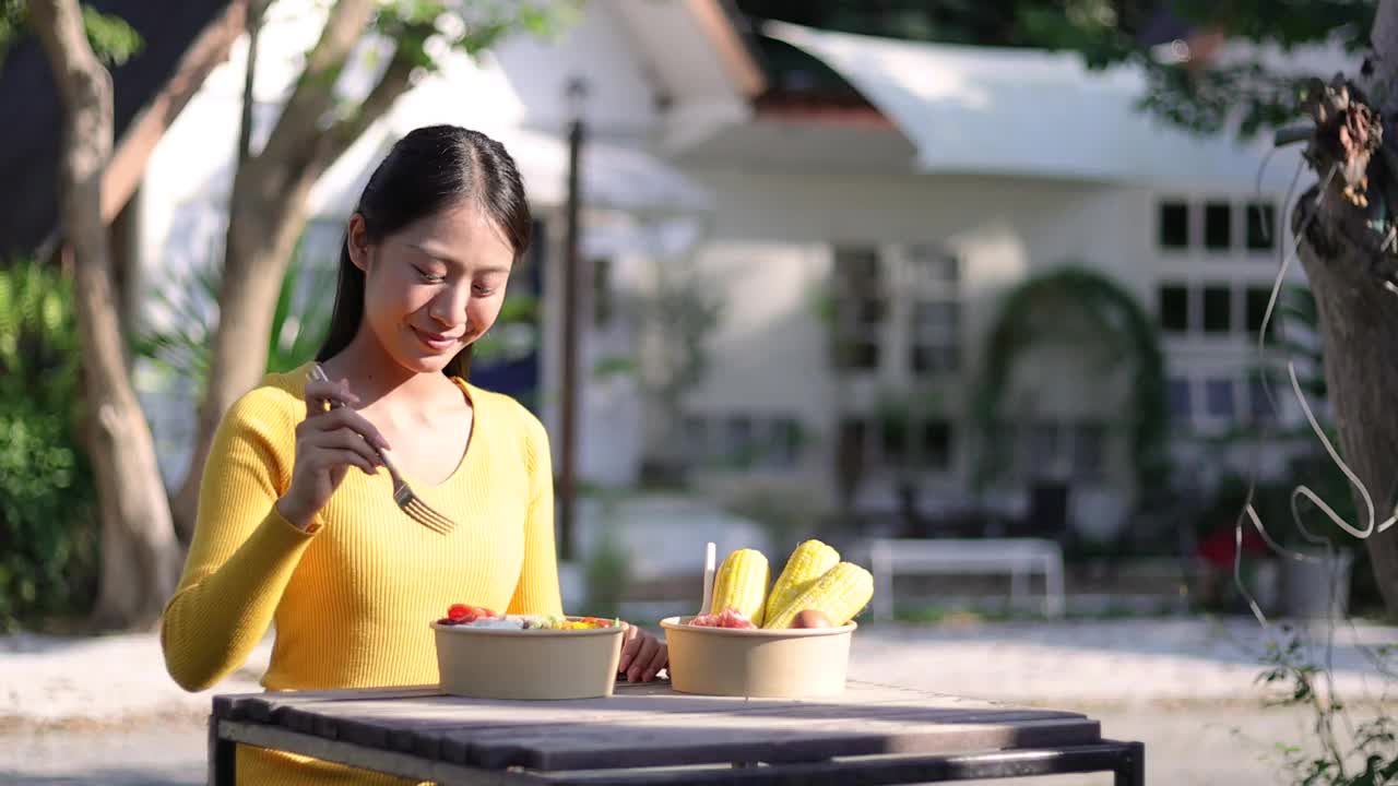 快乐的年轻亚洲女人在后院的木桌上吃蔬菜沙拉。视频素材