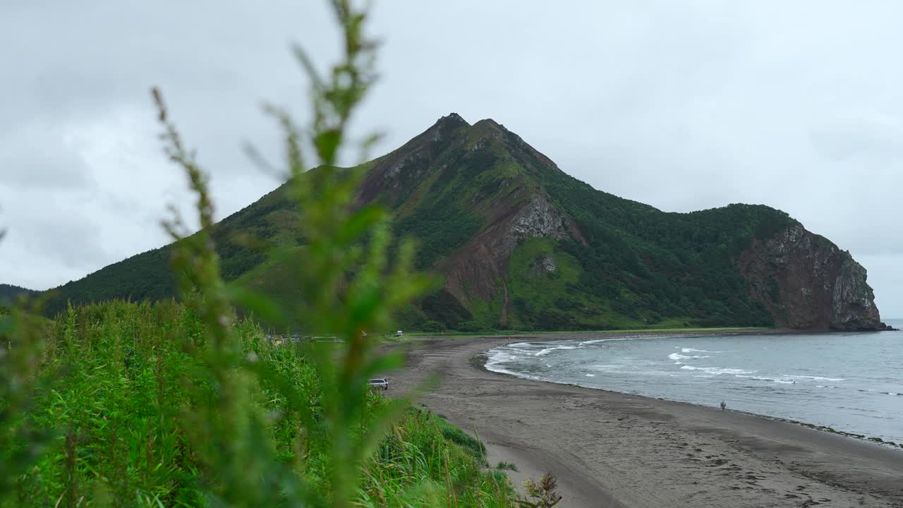 美丽的海滩，绿草和山的景色。夹。风景如画，绿草如茵，以海岸和山为背景。在阴天的海边有沙子和岩石山视频素材