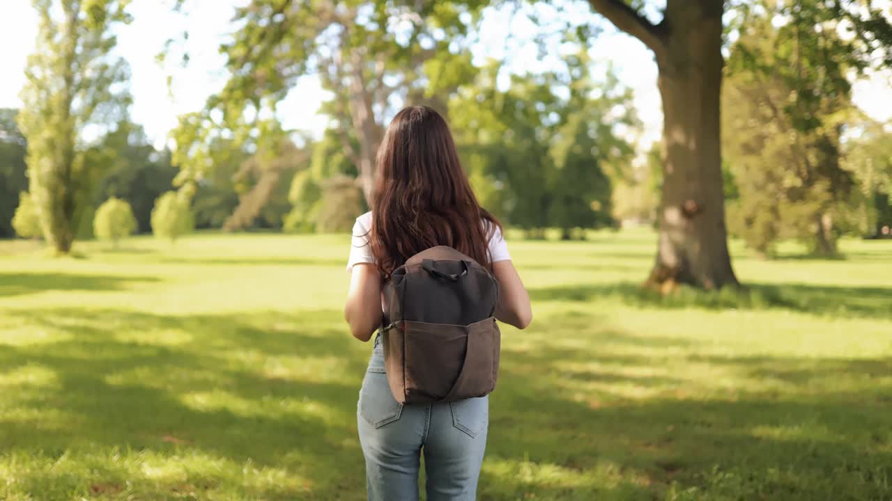 镜头跟随年轻迷人的长发女女孩在夏天的自然公园里背着背包走在户外，转向镜头，笑得很可爱视频下载