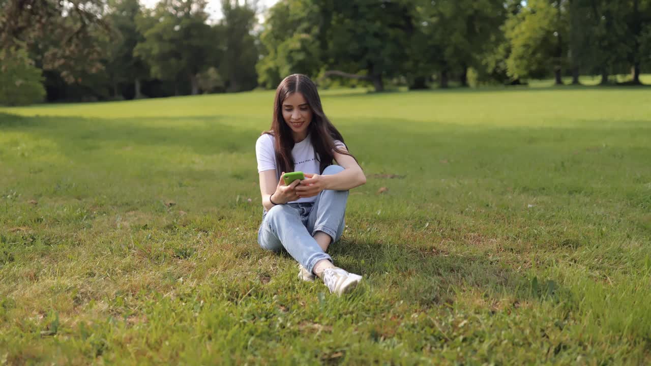 年轻迷人的女学生的肖像，坐在草坪上，在公园的草地上，使用智能手机，微笑，看着相机视频下载