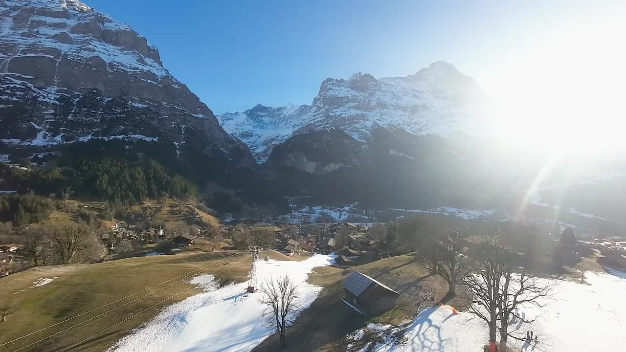 灿烂的阳光照耀着遥远的阿尔卑斯山峰上的格林德沃村。有雪的滑雪场和运动中的缆车。无人机鸟瞰图。视频素材