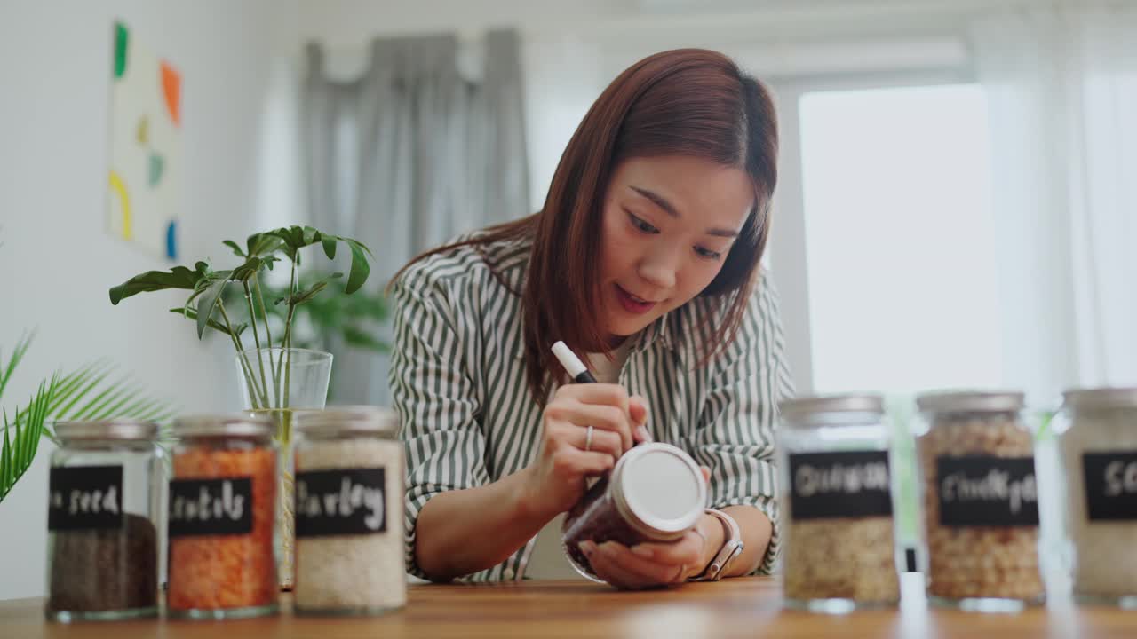 一名亚洲女性用手在木制柜台上的玻璃罐里写着黑色标签的“红豆”，这是一种有组织的厨房食品储藏室，零浪费的生活方式，绿色购物，无塑料和可持续性视频素材