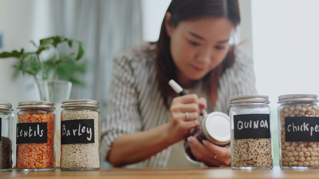 特写:一个女人用手在一个装满厨房食品柜的玻璃罐里写着“红豆”和黑色标签，这是一个有组织的厨房食品柜，零浪费的生活方式，绿色购物，无塑料和家庭可持续发展视频素材