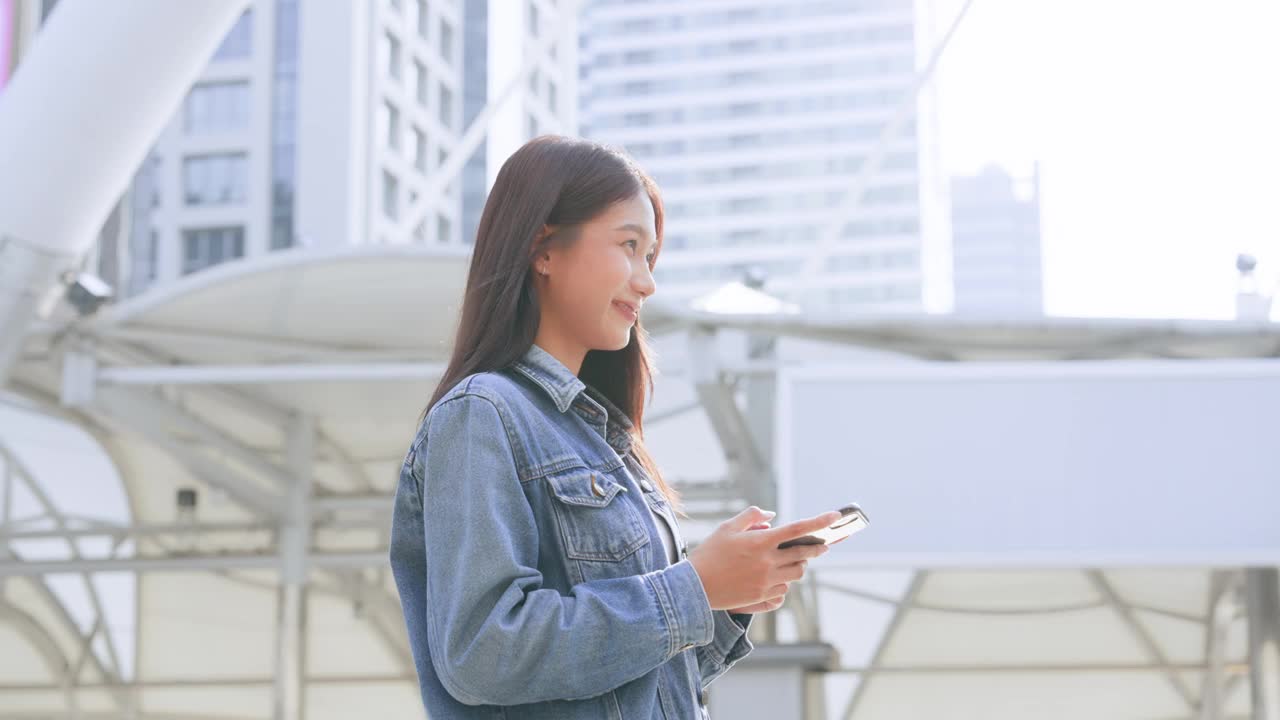 年轻的亚洲女子穿着牛仔裤，夹克，衣服，在晴朗的日子里走在城市的街道上，用智能手机发短信视频素材