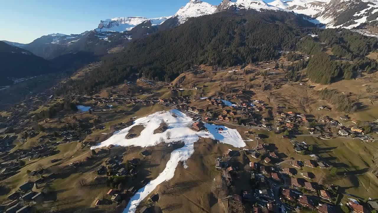 广阔的鸟瞰图的村庄在阿尔卑斯山滑雪坡覆盖在绿色山谷的中心积雪。冬末的瑞士格林德沃。视频素材