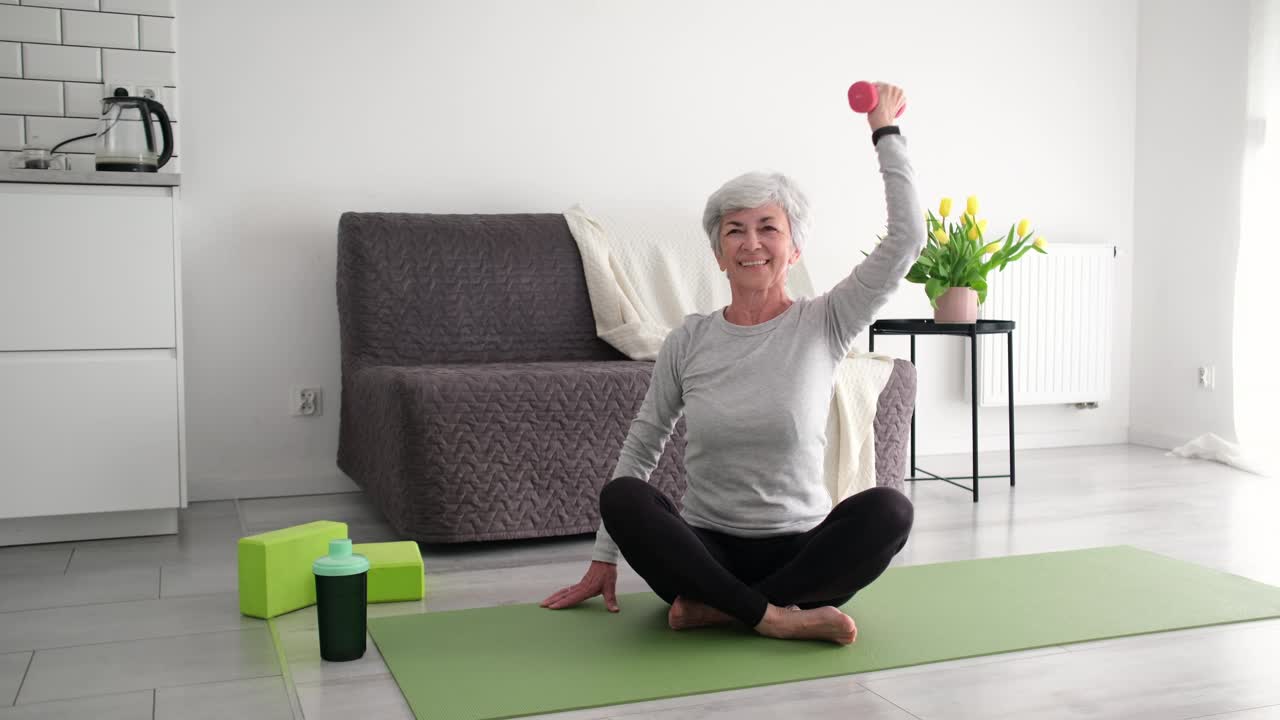 微笑的老年妇女在家里的瑜伽垫上用哑铃做运动视频下载