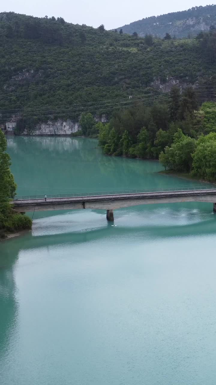 无人机俯瞰绿松石山湖和横跨湖面的桥梁。翡翠湖坐落在岩石地形和茂密的森林中。桥跨湖，展现宁静的湖、桥和周围的自然视频素材