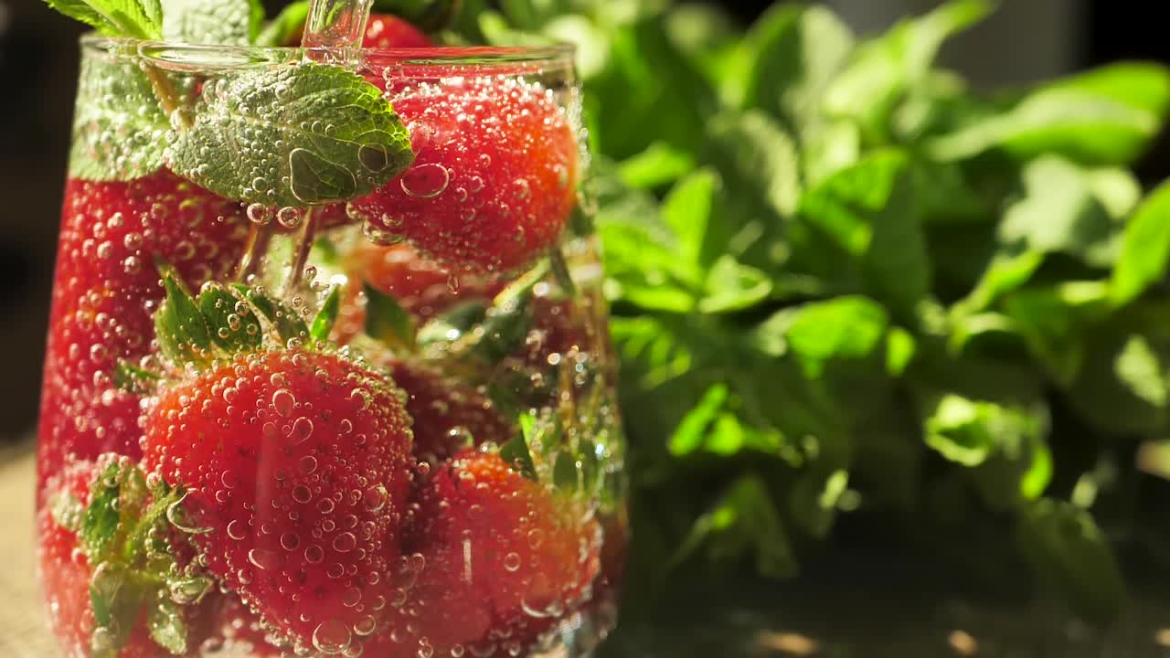 一个杯子的特写，里面装满了苏打水、大的新鲜草莓和带有大气泡的绿薄荷叶。清爽的浆果碳酸饮料。可重复使用的玻璃吸管。本空间视频素材