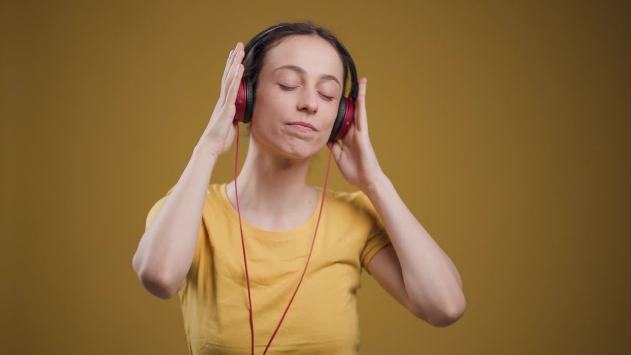 一个安静的年轻女子通过红色耳机听音乐，闭上眼睛，在鲜艳的黄色背景下温柔地微笑。视频素材