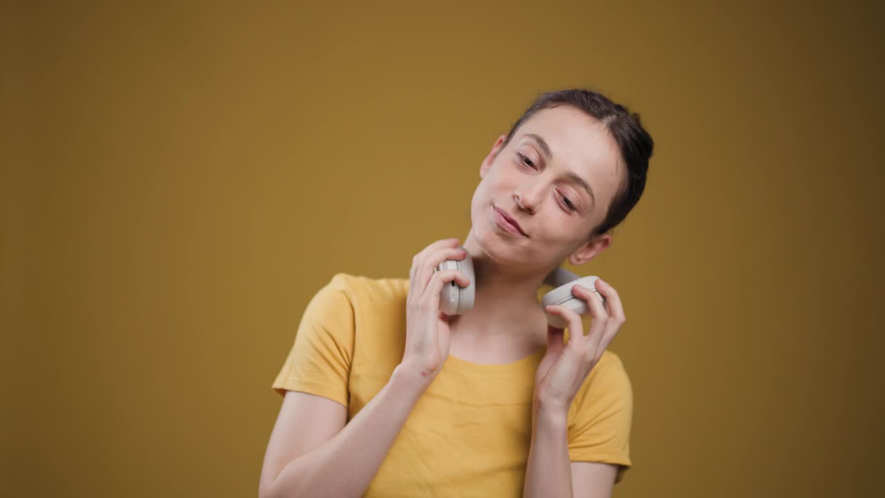 这张迷人的照片中，一位快乐的年轻女子戴着耳机，开心地微笑着，在诱人的黄色背景下听着她最喜欢的音乐。视频素材