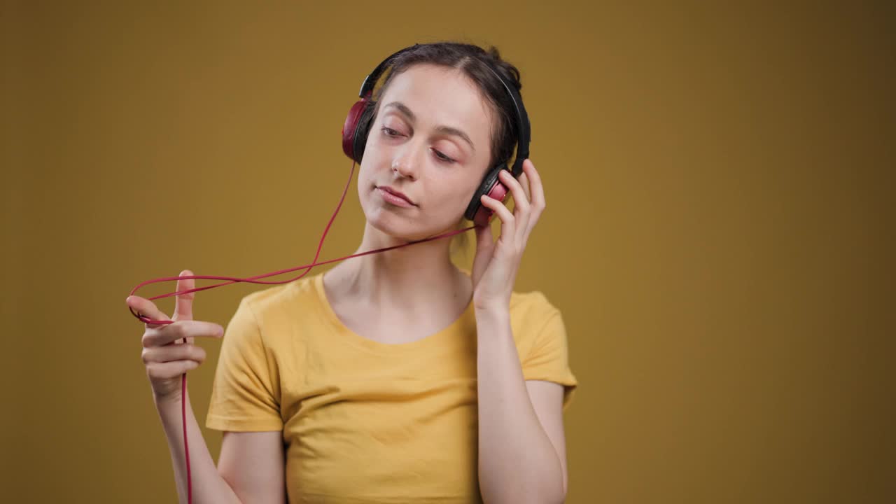 一个安静的年轻成年女子闭上眼睛，在普通的黄色背景下通过红色耳机听音乐。视频素材