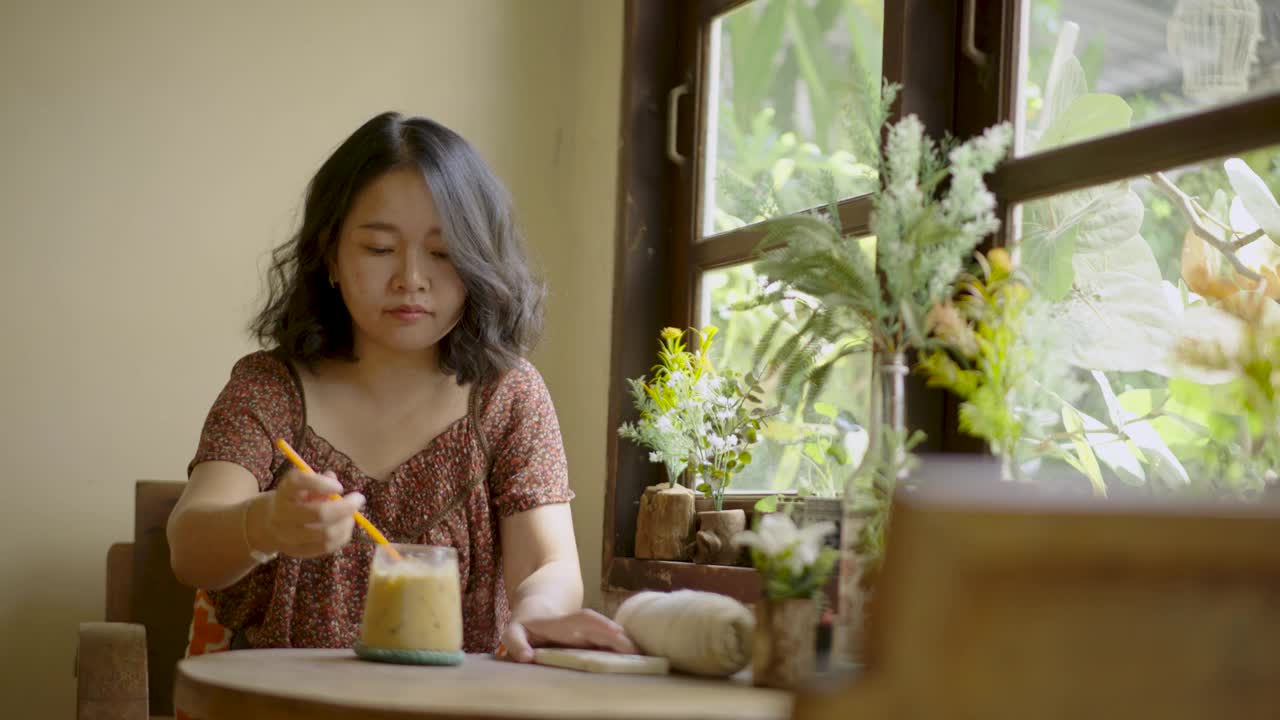 迷人的亚洲年轻女子在咖啡馆阳光明媚的靠窗座位上享受咖啡。视频下载