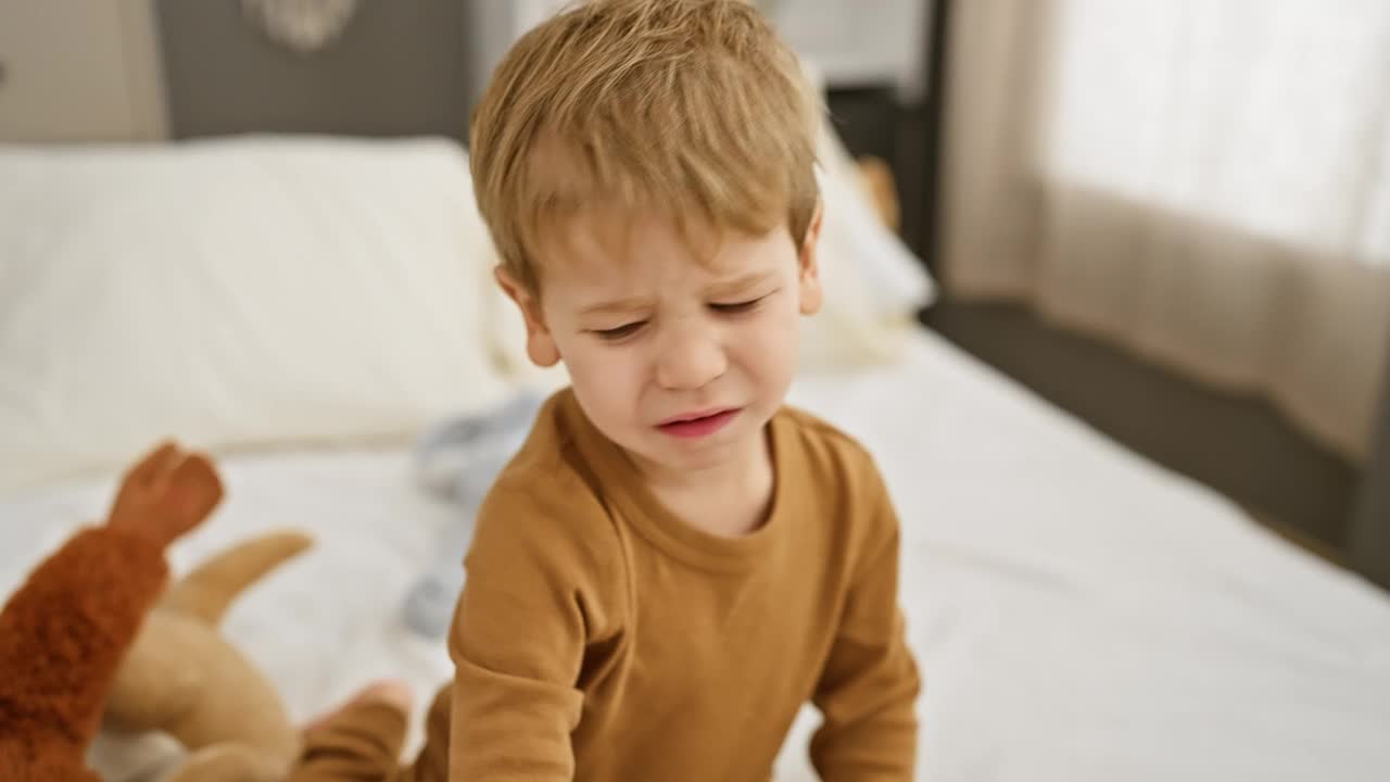 一个蹒跚学步的男孩穿着米色的衣服，在室内的白色床上玩毛绒玩具，唤起了天真和童年。视频素材