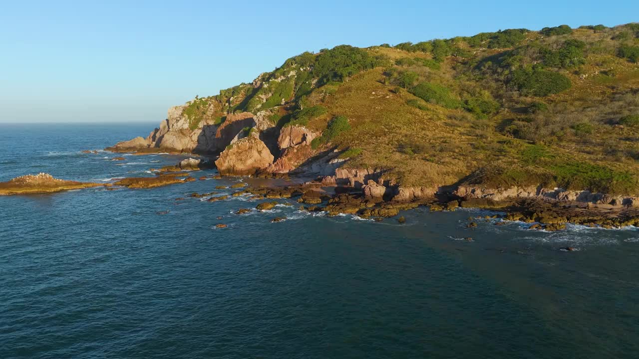 日出时一个荒岛的岩石海岸的航拍视频，展示了橙色的岩石被太阳照亮，强大的海浪冲击着海岸，创造了一个戏剧性的海景视频素材