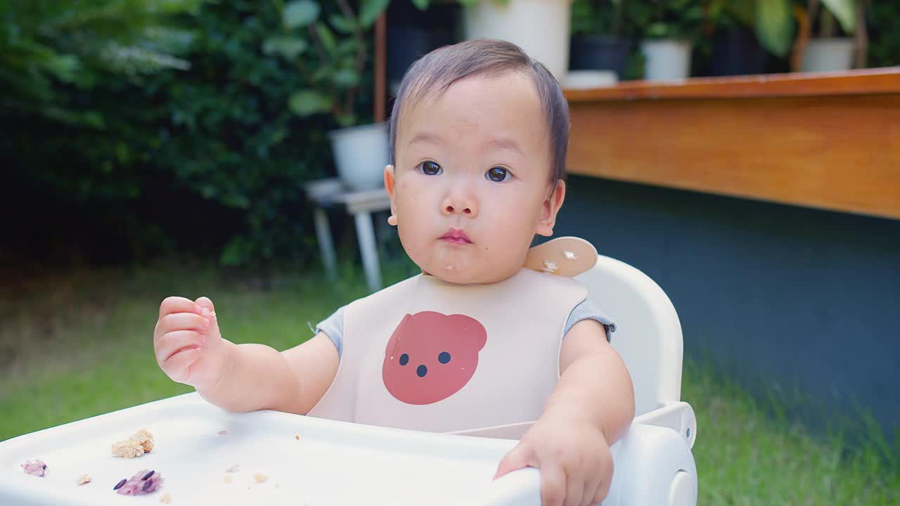 一个婴儿坐在高椅子上，围嘴上戴着一只熊。幼儿用餐时间在公园外面吃食物视频素材