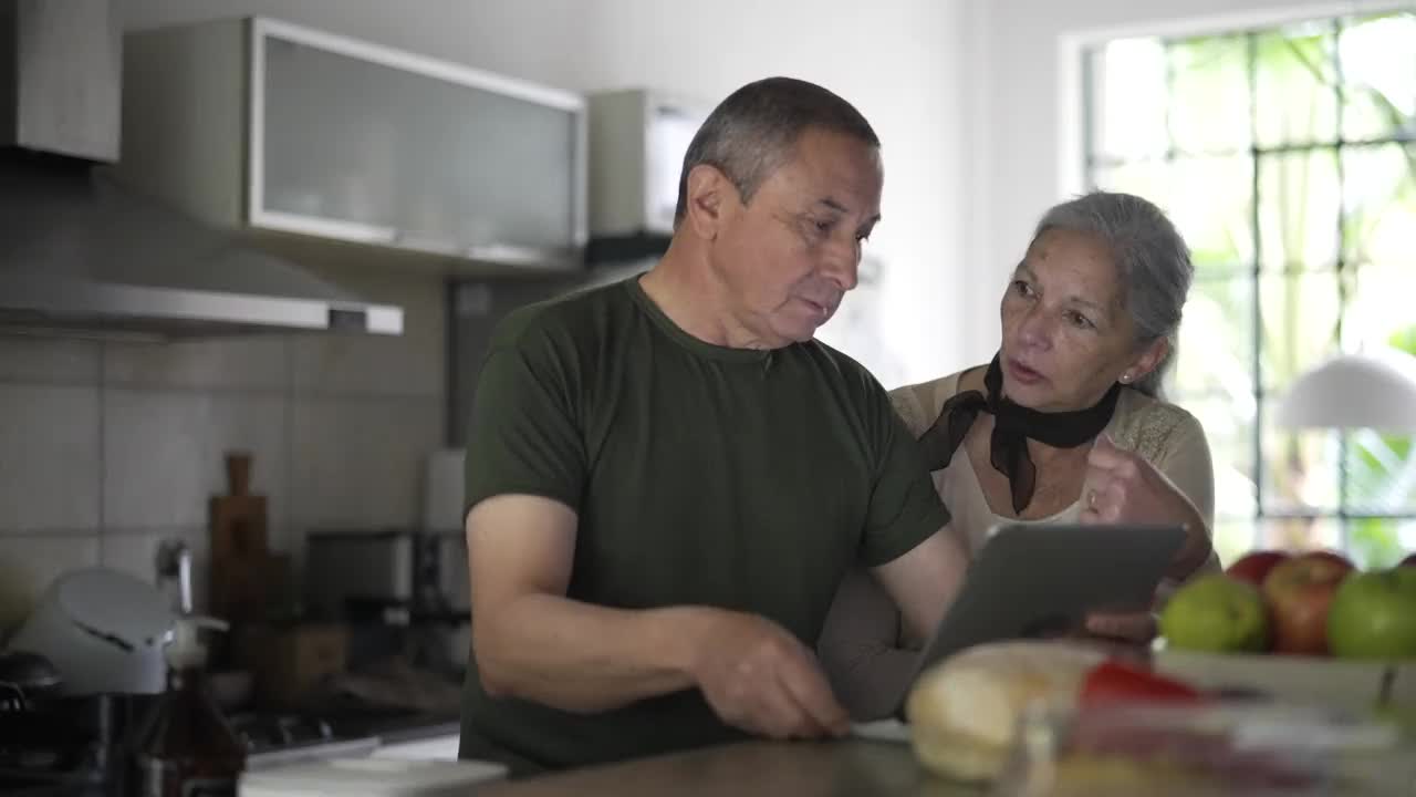 老夫妇一边在厨房里分享时光，一边看着平板电脑上的家庭照片视频素材