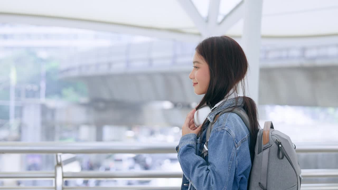 年轻的亚洲女子穿着牛仔夹克，脖子上戴着耳机，背着背包走在城市里。青春期的女性开心，笑脸盈盈。放松和享受城市生活的年轻女士视频素材