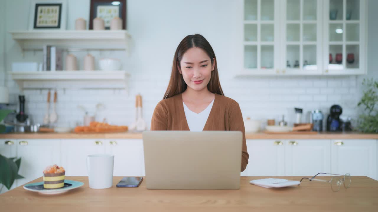 年轻漂亮的亚洲女人在家里厨房用笔记本电脑工作。在家工作视频下载