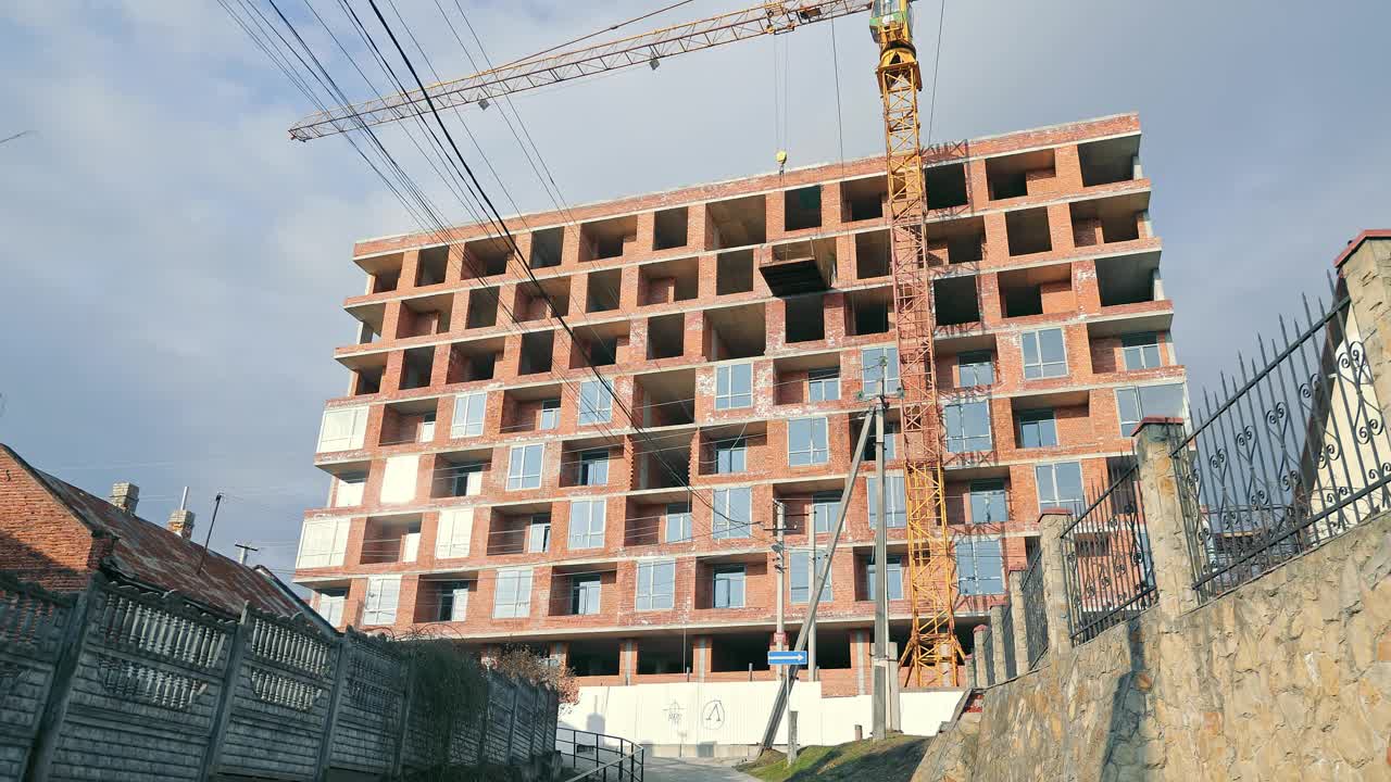 正在开发的现代城市建筑工地，在城市环境中建造的大型多层建筑，以高耸的起重机和未完工的砖立面为特色，沐浴在阳光下。视频素材