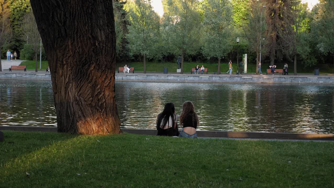傍晚时分，两个年轻女子坐在一个小湖边的草地上。视频下载