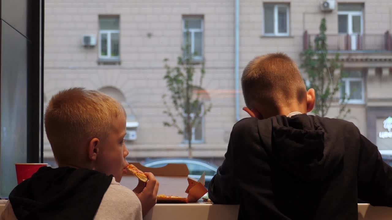 两个男孩坐在窗前的高木桌上吃披萨。视频下载