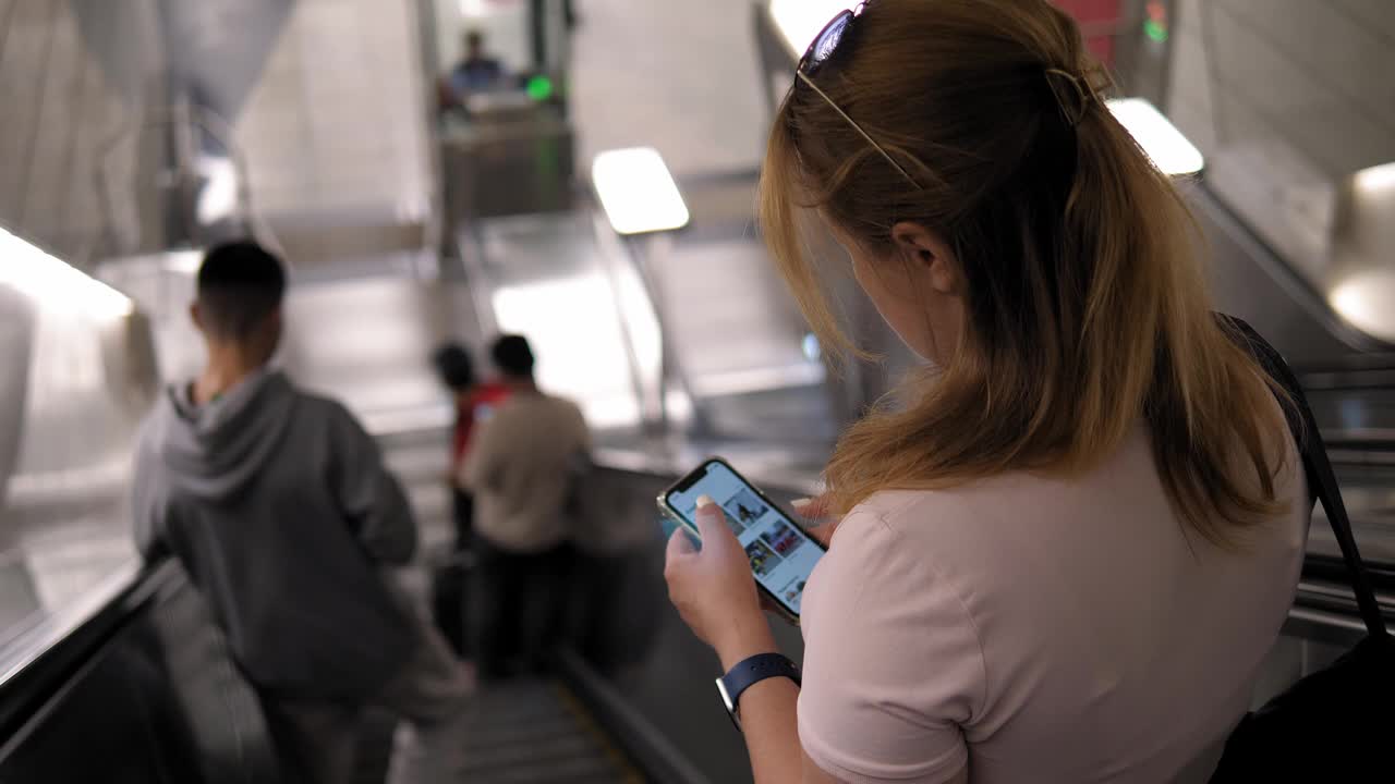 在地铁的自动扶梯上，一位手持手机的年轻女子从背后看到的景象视频下载
