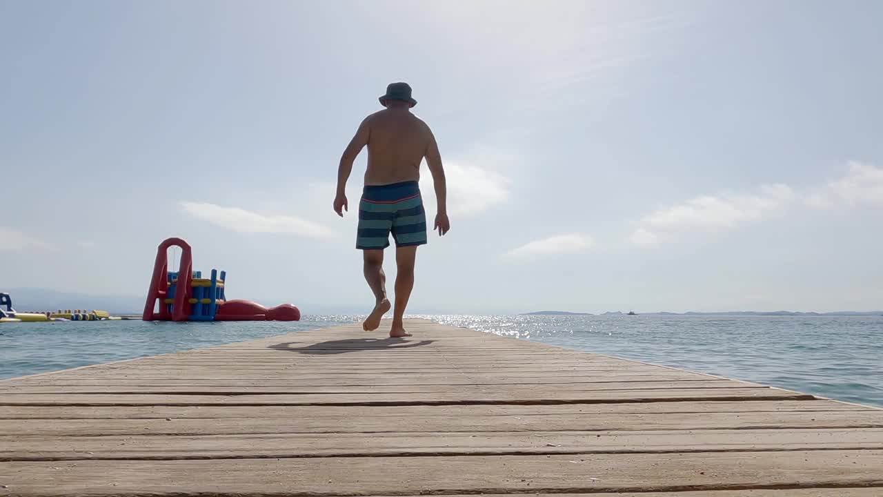 一个穿着短裤、戴着帽子的男人慢镜头走过这个热带岛屿上的一个木制码头视频素材