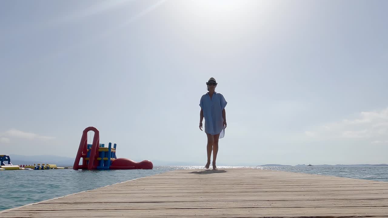 年轻的黑发女子戴着帽子走在沙滩上的木制码头上，在暑假，慢动作视频素材