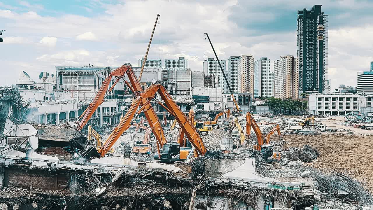 这是一个熙熙攘攘的城市拆迁现场，在部分被毁建筑的废墟中，多台挖掘机和起重机在高层建筑和多云的天空的背景下，展示了城市改造和控制的动态过程视频下载