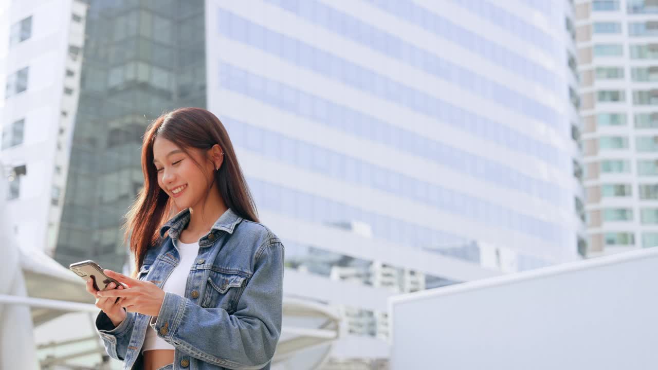 年轻的亚洲女子穿着牛仔裤，夹克，衣服，在晴朗的日子里走在城市的街道上，用智能手机发短信视频素材