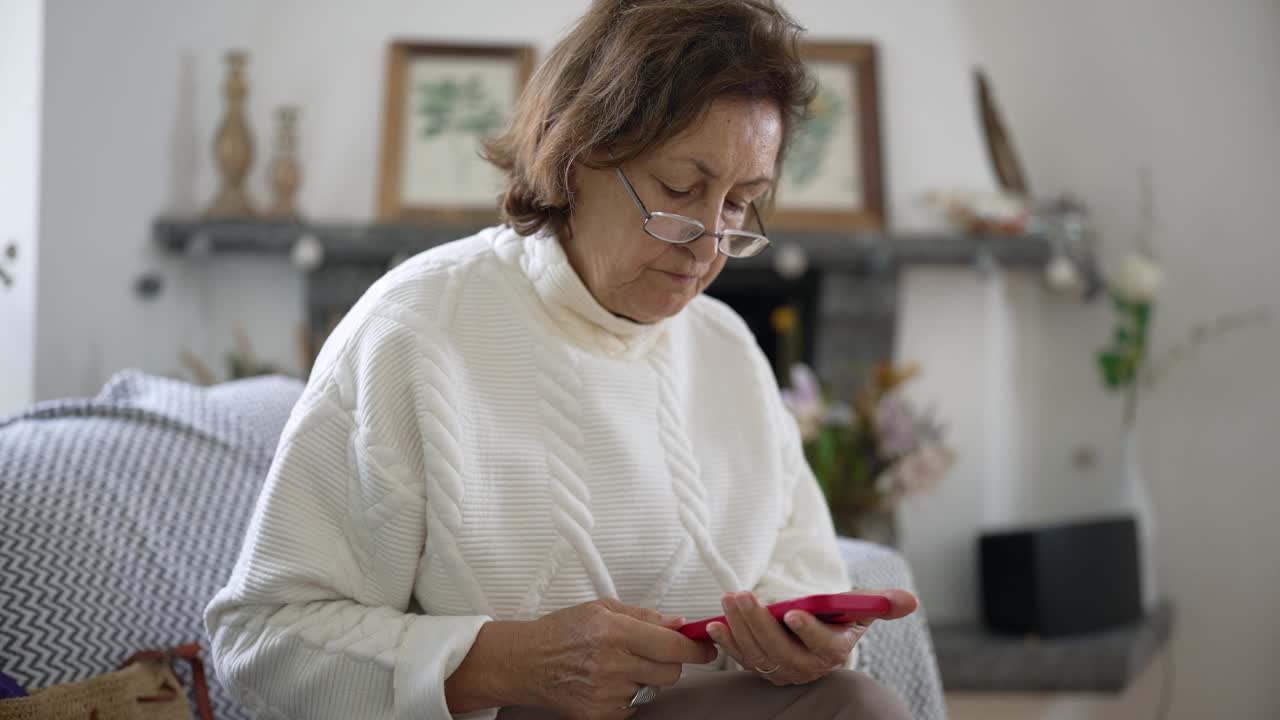 一位高级女性全神贯注地使用智能手机，说明她积极参与科技活动，舒适地坐在装饰精美、气氛舒适的客厅里视频素材