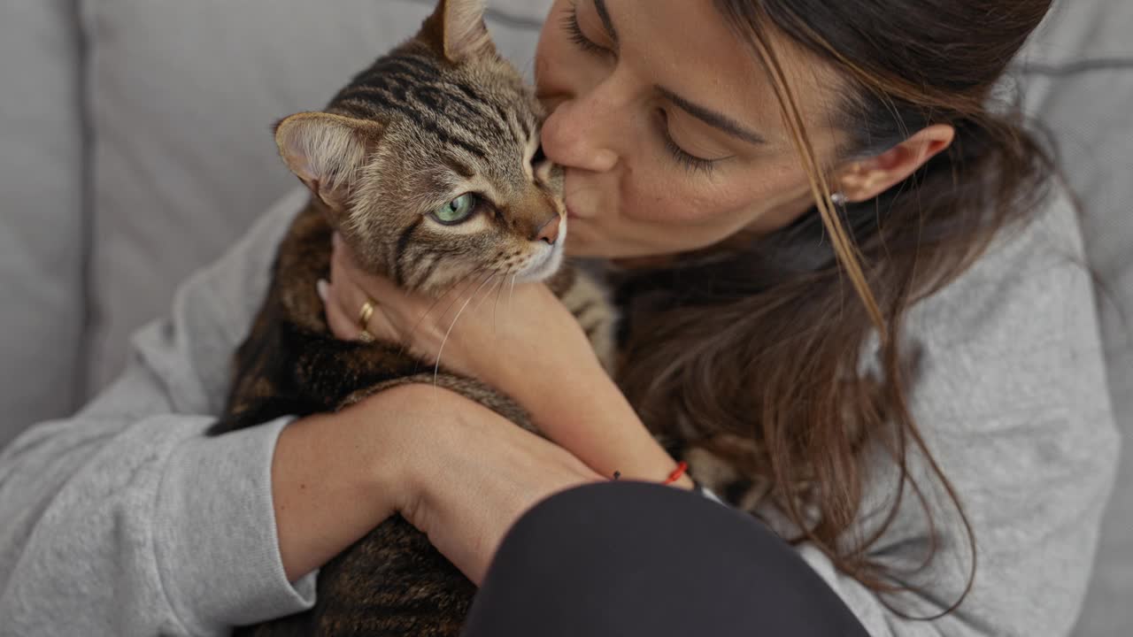 一位年轻的西班牙妇女在舒适的客厅里亲吻并拥抱她的虎斑猫，表达爱意和陪伴。视频素材